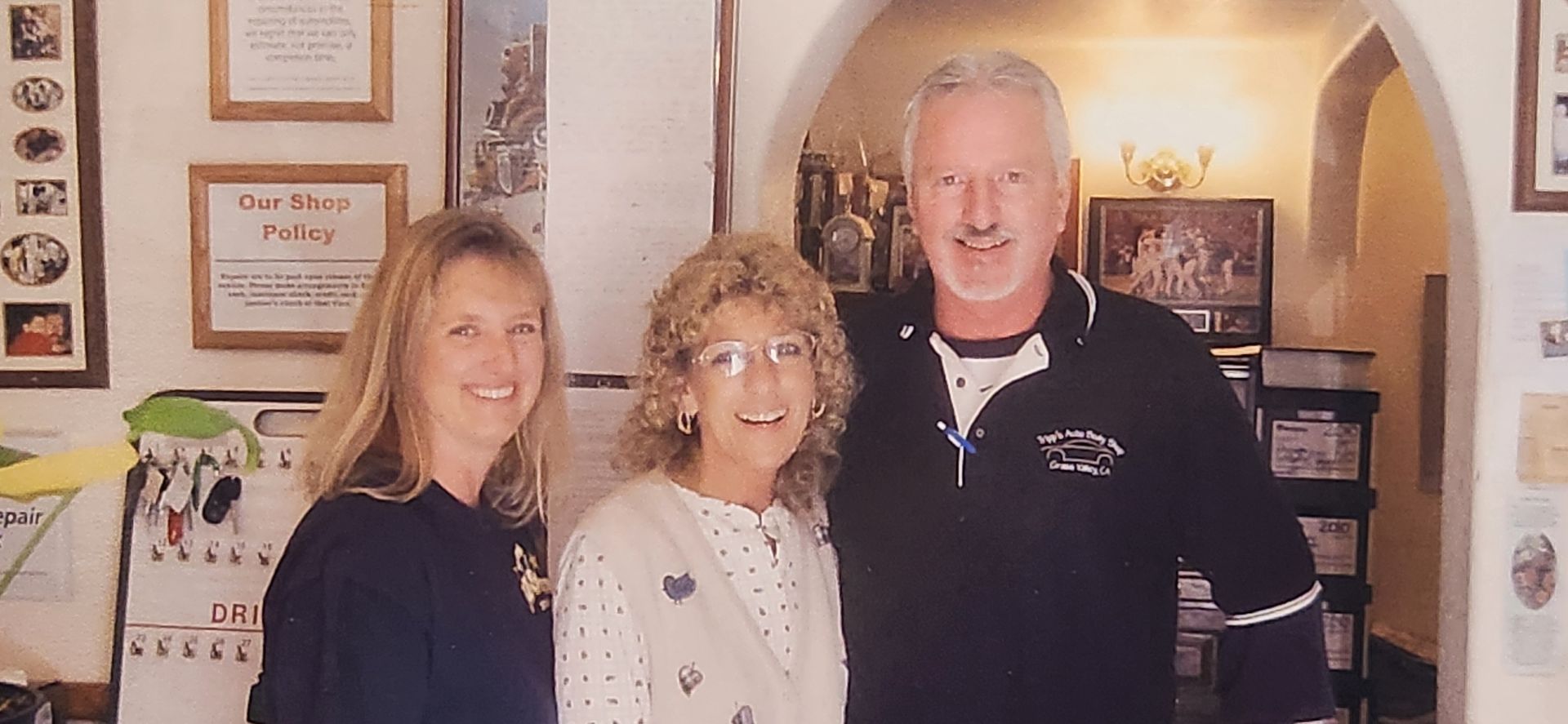 Three people smiling indoors in front of a display wall and arched doorway. | Tripp's Auto Body