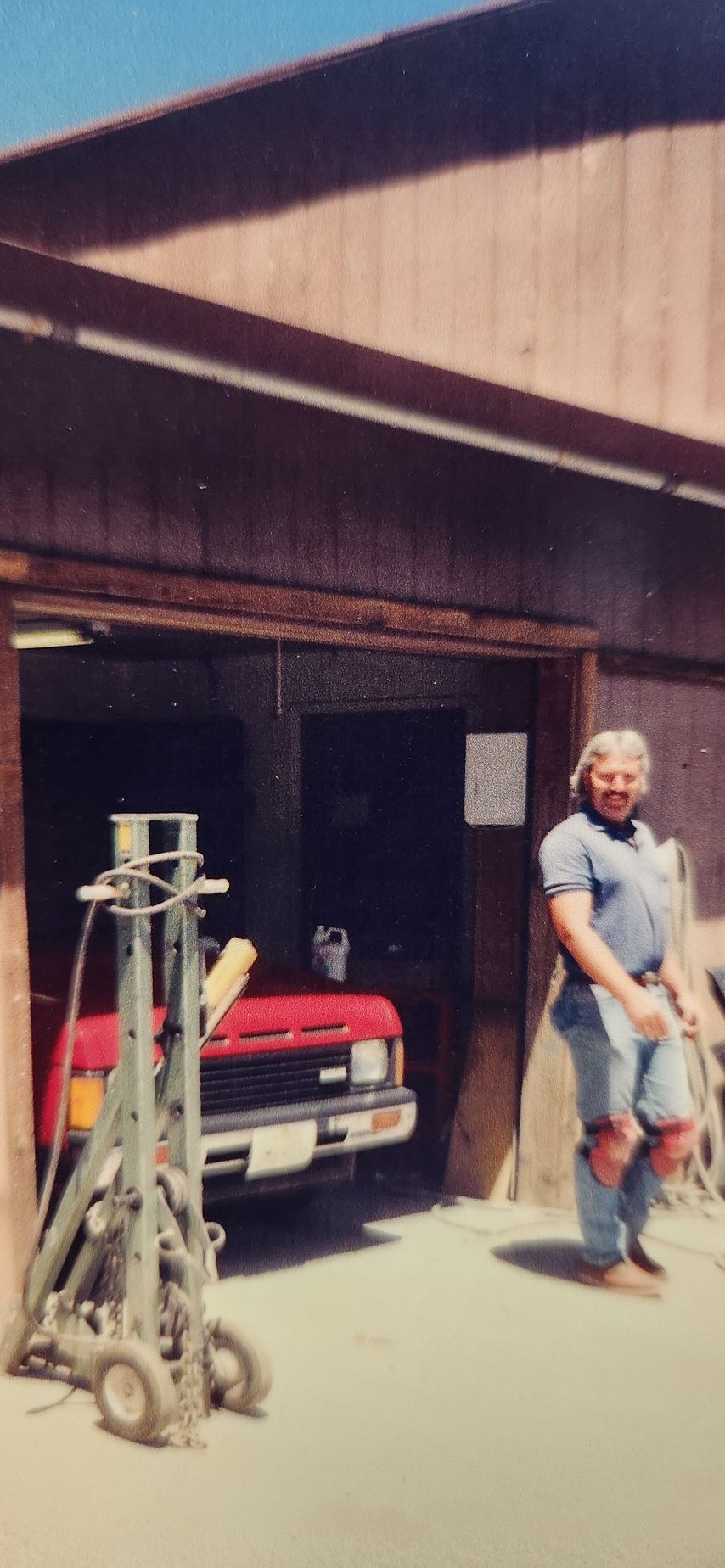 Man standing outside a garage beside a red car and stacked tools in bright sunlight | Tripp's Auto Body