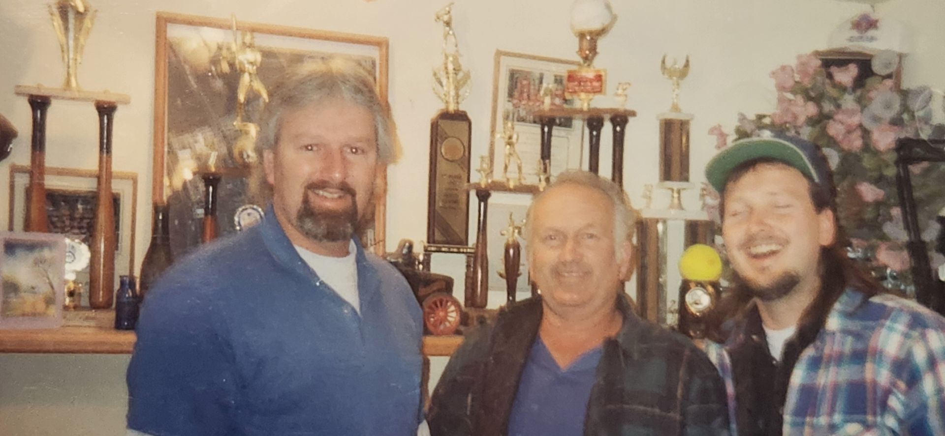 Three men smiling in a room with trophies, framed art, and floral decor. | Tripp's Auto Body