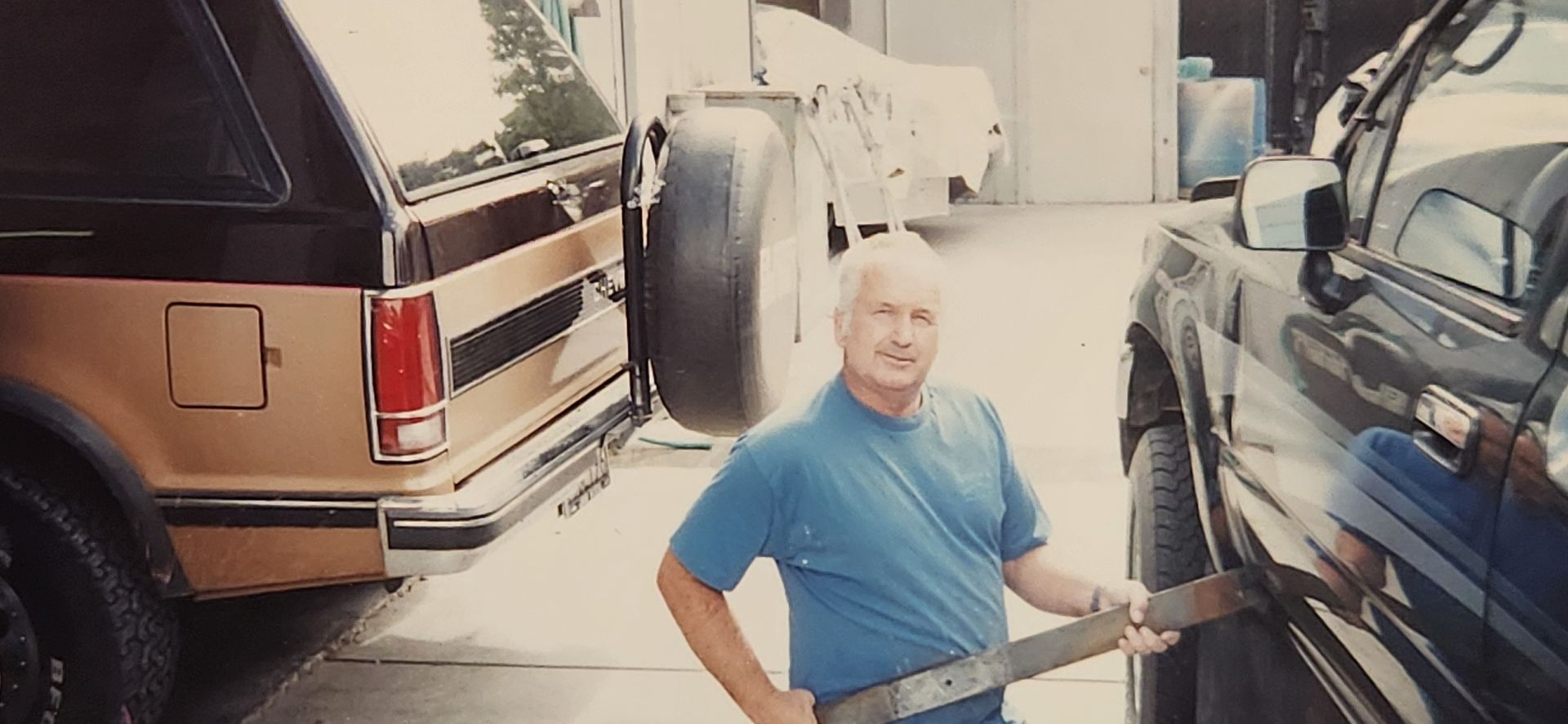 Man in blue shirt standing between two parked vehicles in a driveway, holding a long tool. | Tripp's Auto Body