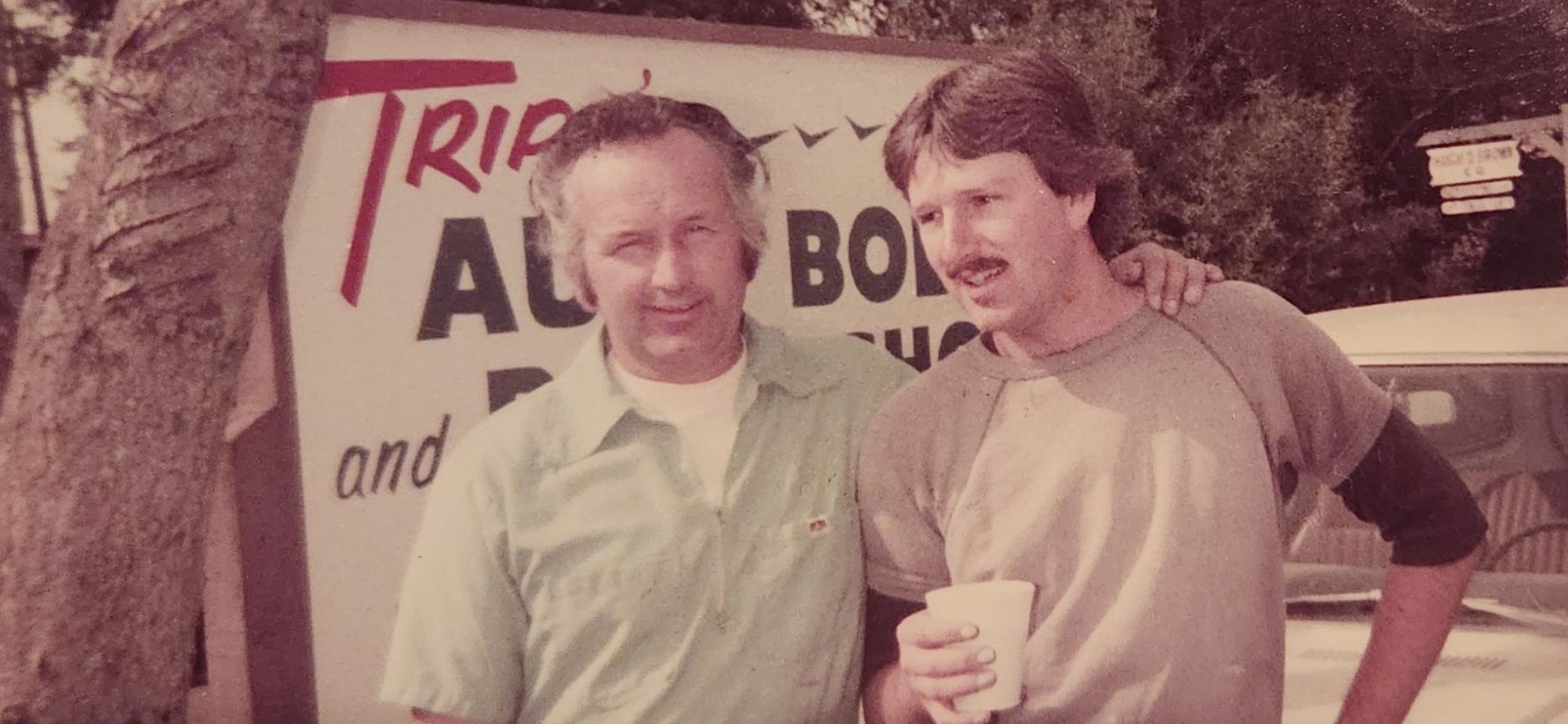 Two men standing outdoors in front of a sign, one holding a cup and smiling. | Tripp's Auto Body
