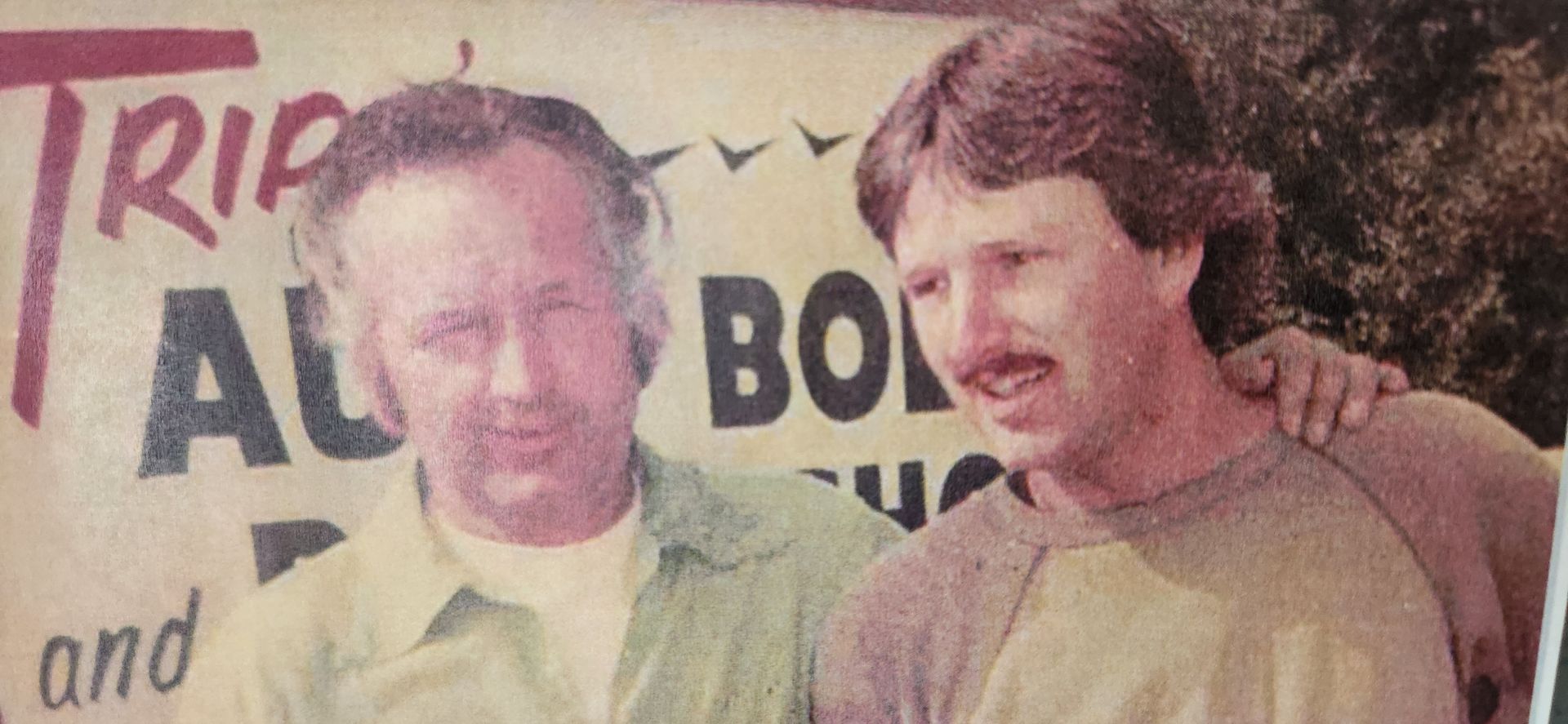 Two men standing together in front of a sign, one with a hand on the other's shoulder. | Tripp's Auto Body