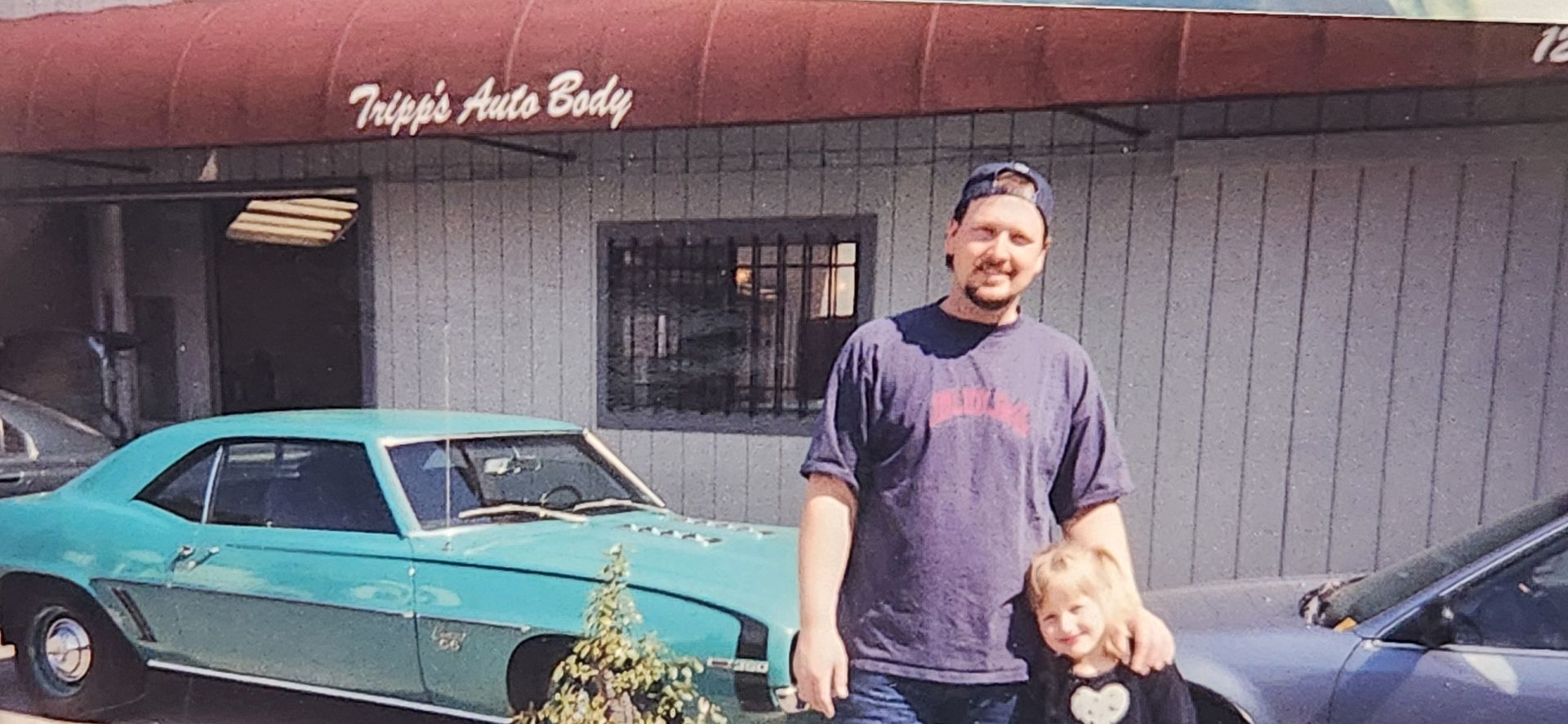 Man and child standing beside a turquoise classic car in front of a storefront. | Tripp's Auto Body