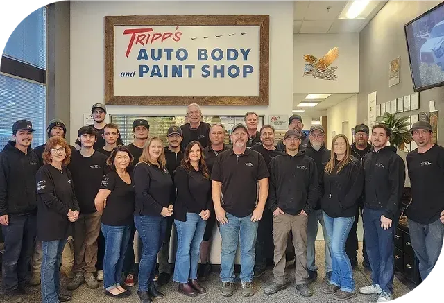 Group of auto body shop staff posing indoors in front of a Tripp’s Auto Body and Paint Shop sign | Tripp's Auto Body
