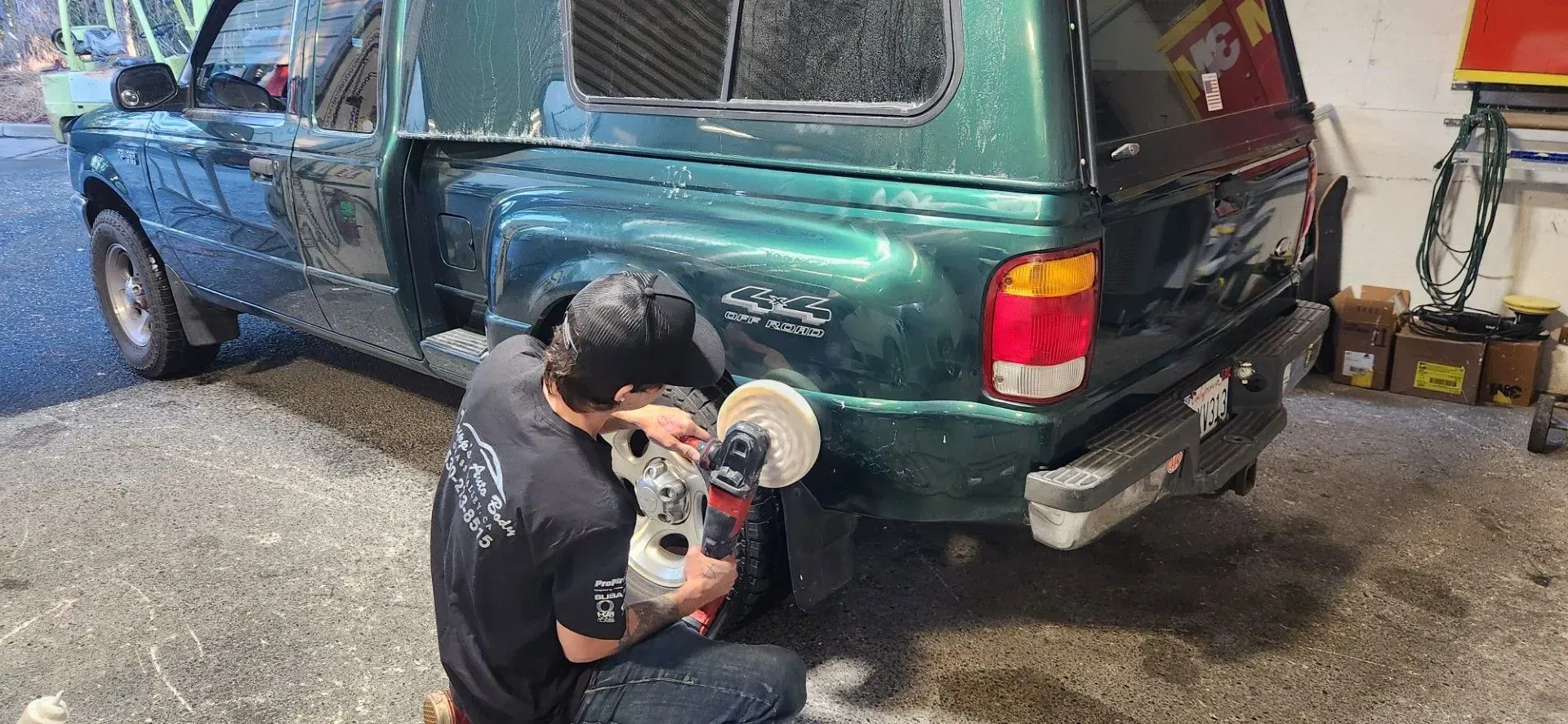 Person crouching by a green SUV, using a machine on the rear quarter panel in a garage. | Tripp's Auto Body