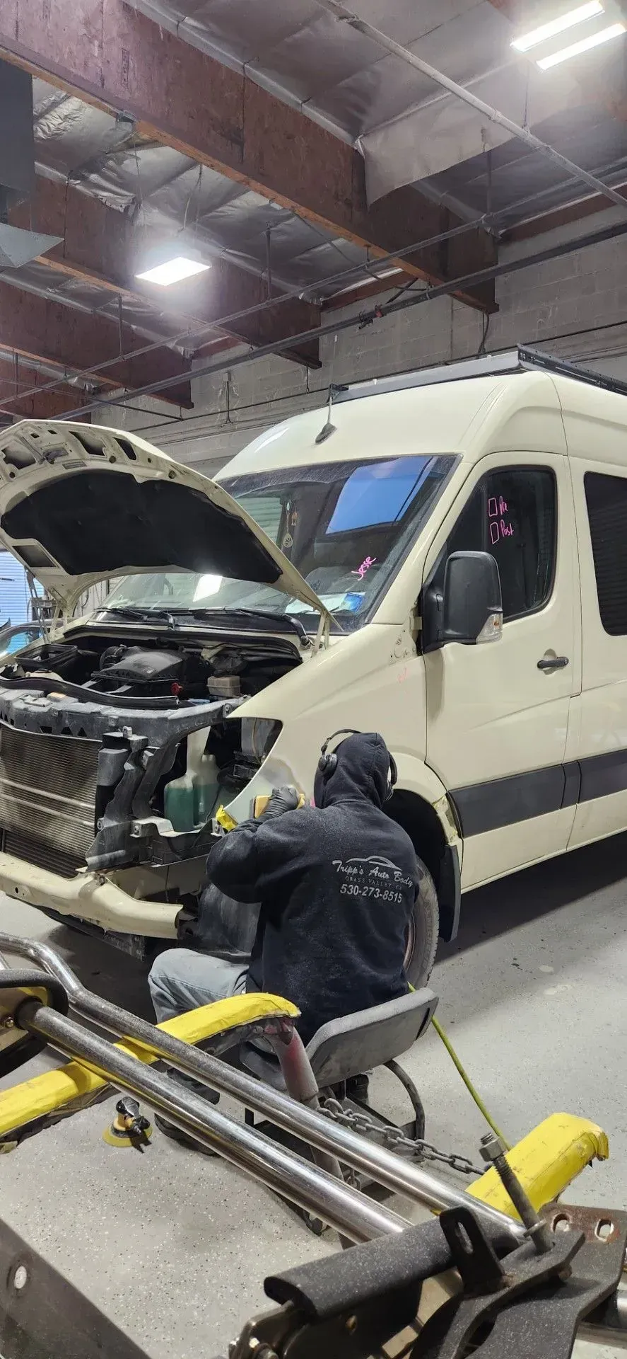 Person working on a white van with open hood in a garage, using a metal cart and tools | Tripp's Auto Body