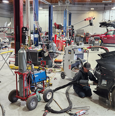 Auto repair shop with tools and lifts; mechanic works on a car near the front bumper. | Tripp's Auto Body