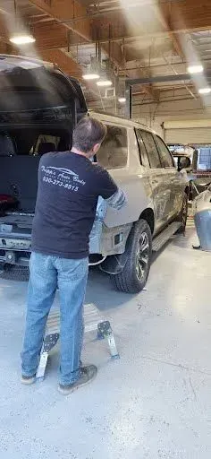 Two men working on a white SUV in a workshop, one using a tool near the rear wheel. | Tripp's Auto Body