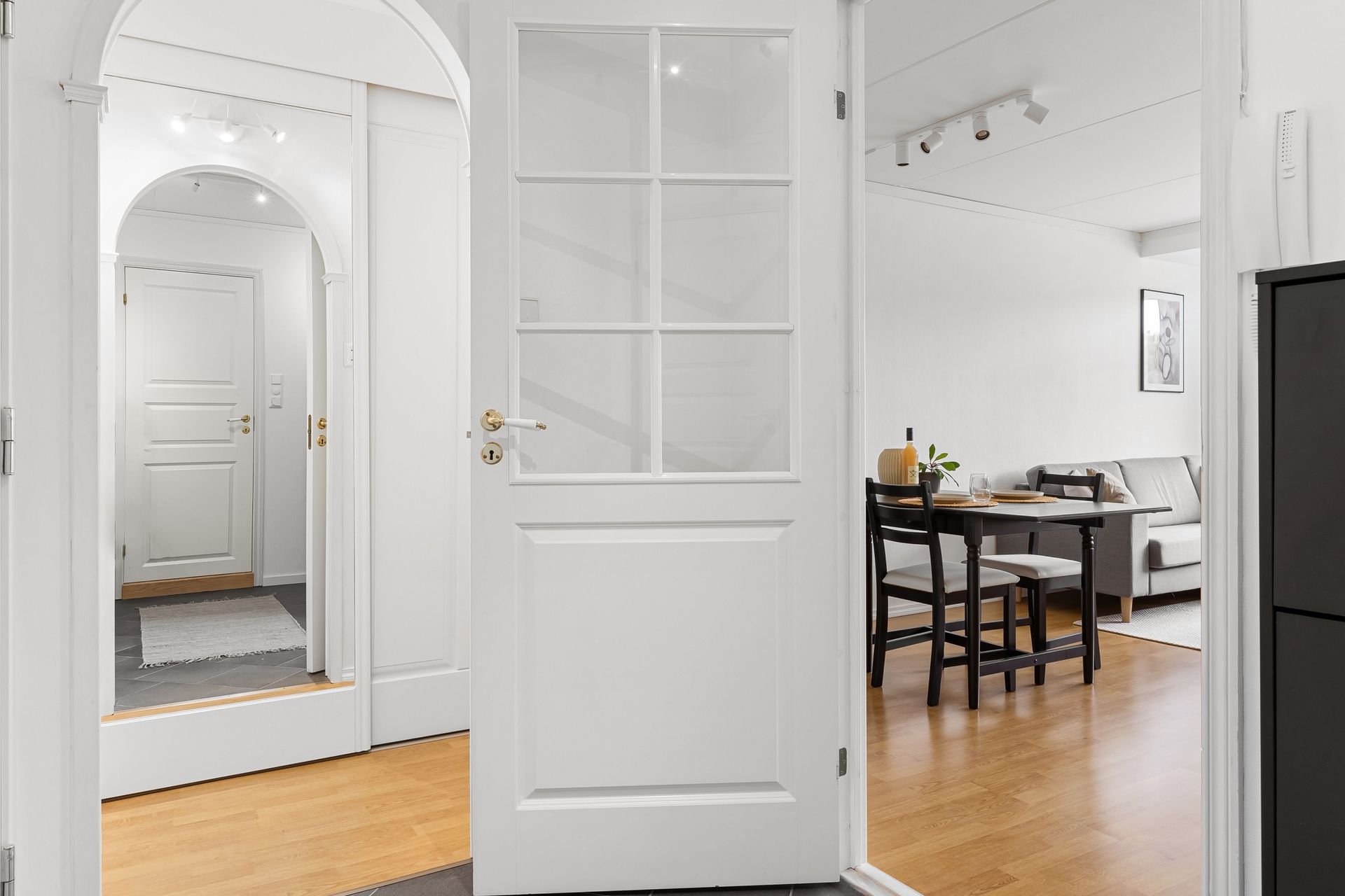 Bright white apartment hallway with a mirror, open door, and dining area with table and chairs.