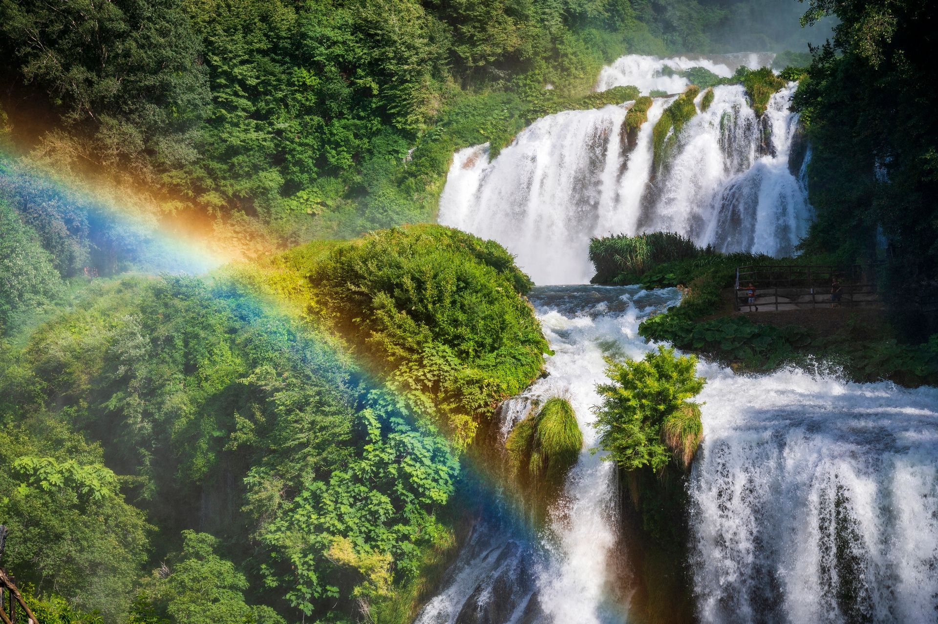 Una lussureggiante cascata che scende a cascata sulle rocce, con un arcobaleno nella nebbia, circondata da una fitta foresta verde.