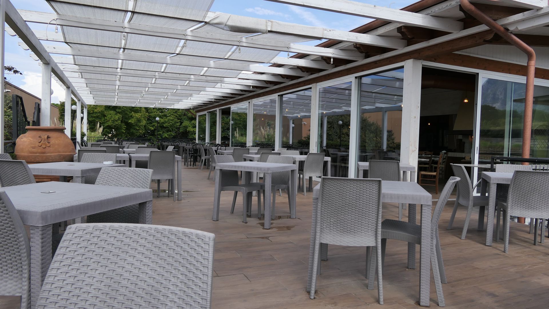 Terrazza esterna vuota del bar, con tavoli e sedie sotto una tettoia a pergola.