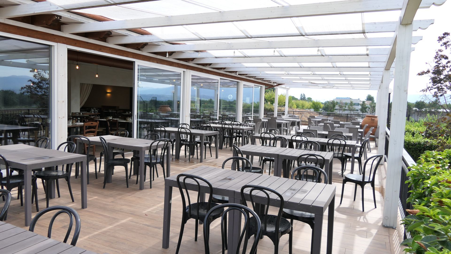 Patio esterno con tavoli e sedie neri sotto una tettoia bianca, con vista panoramica.
