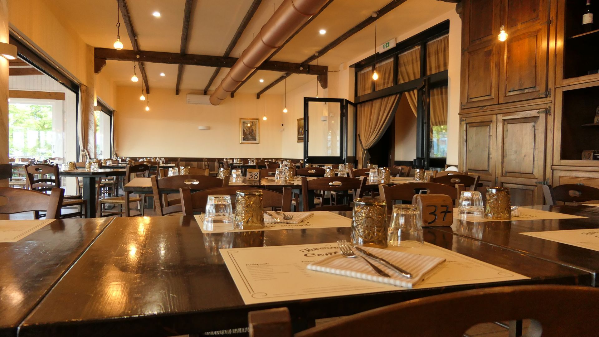 Sala da pranzo vuota di un ristorante moderno con tavoli e sedie in legno e lampade a sospensione dai colori caldi.