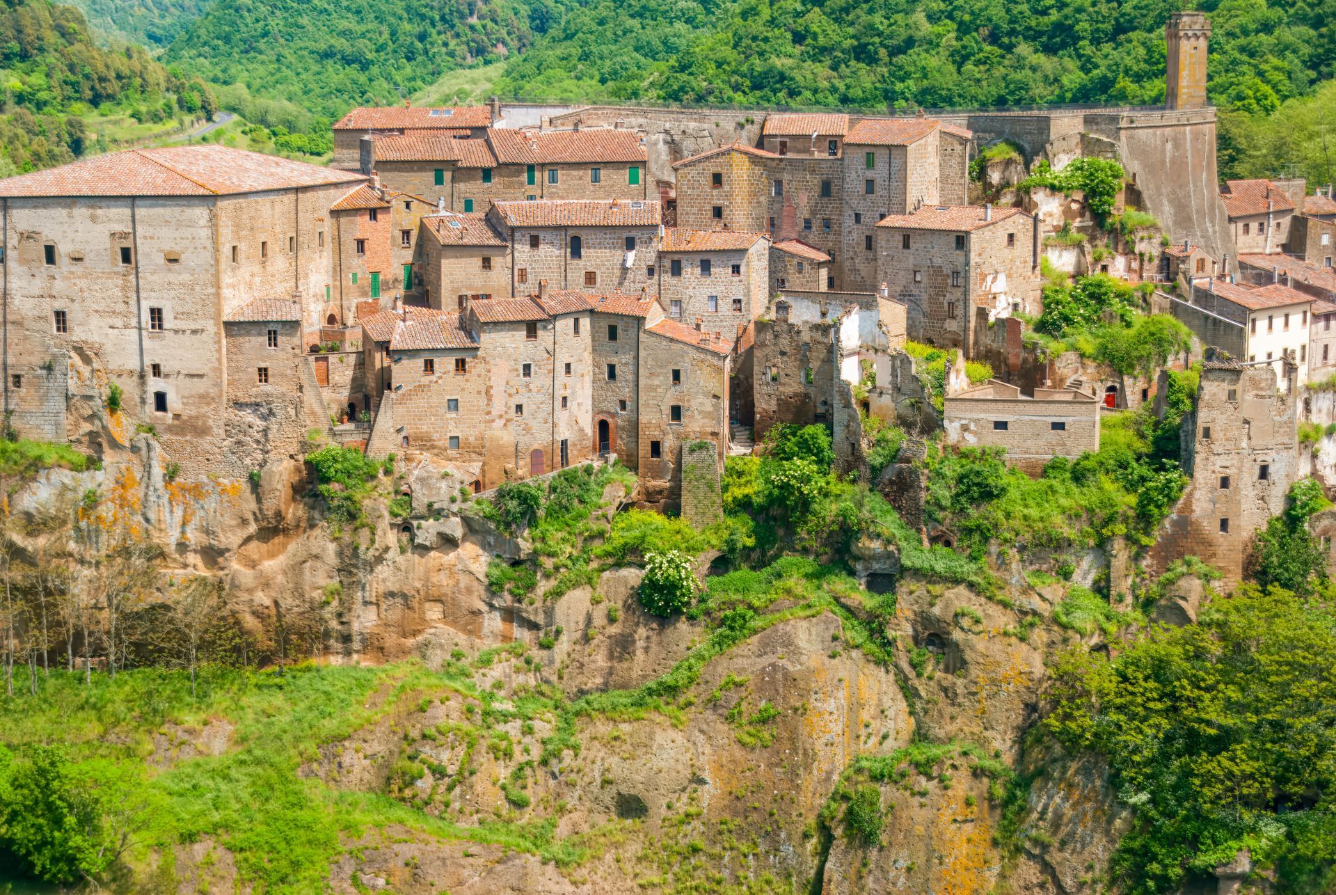Villaggio collinare di edifici in pietra e tetti di terracotta immerso tra verdi scogliere e alberi.