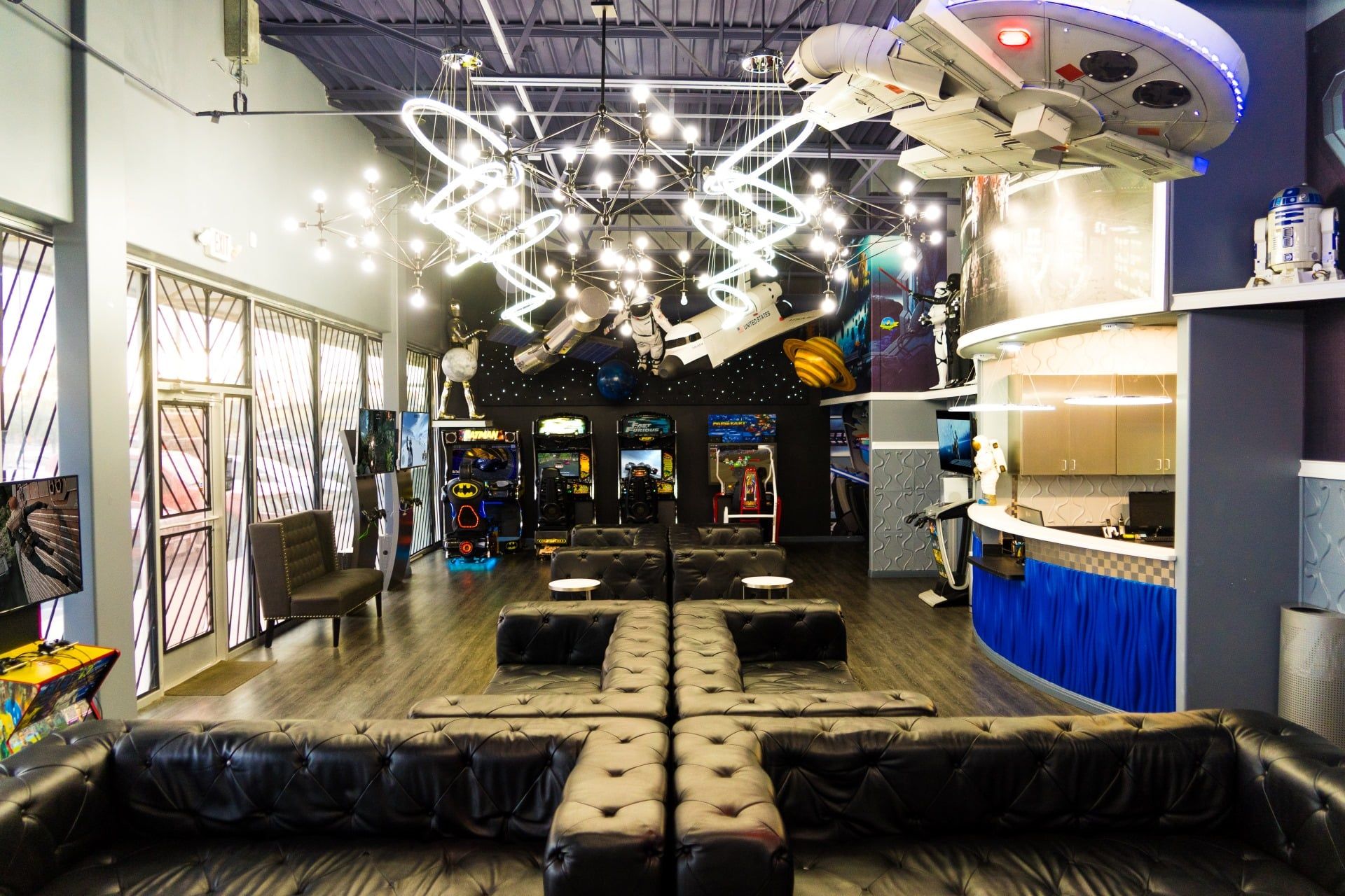 Bright lounge interior with black leather sofas, neon ceiling lights, and arcade machines in back.