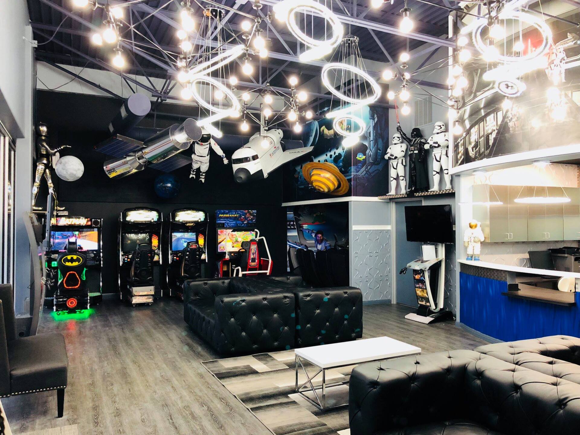 Modern arcade lounge with neon ceiling lights, black tufted seating, and rows of game cabinets