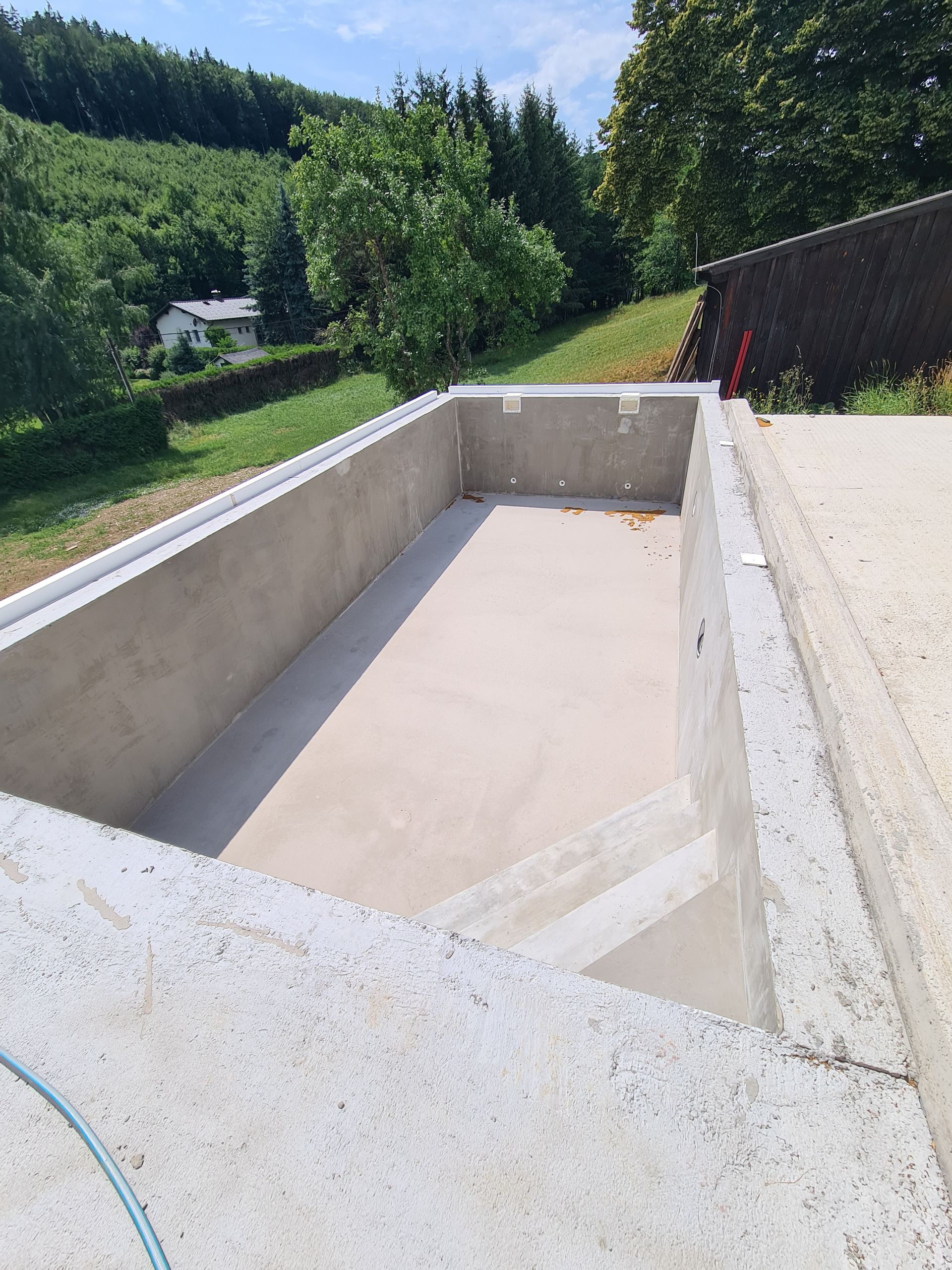 Leeres rechteckiges Betonbecken im Bau, mit einem Grashügel und Bäumen im Hintergrund.