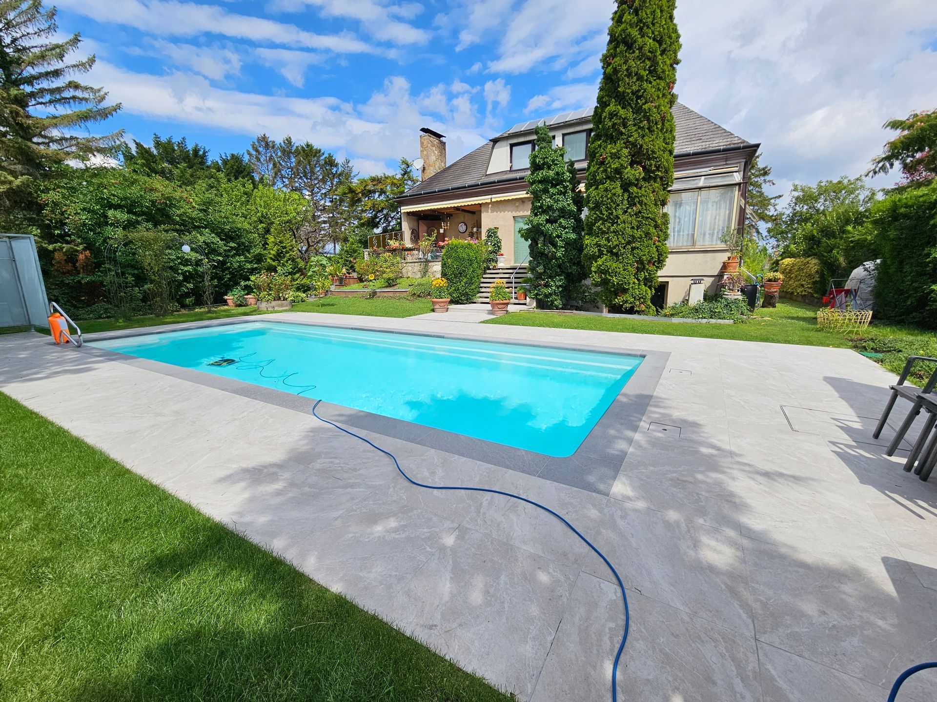 Pool mit blauem Wasser, umgeben von einer Betonterrasse und grünem Rasen. Im Hintergrund ist ein Haus zu sehen.