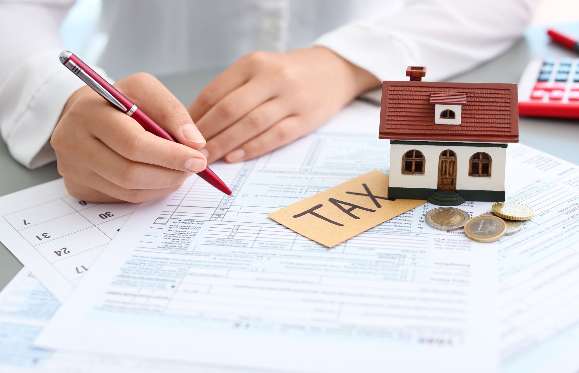 Person writing on tax forms next to a toy house and coins, symbolizing home property taxes