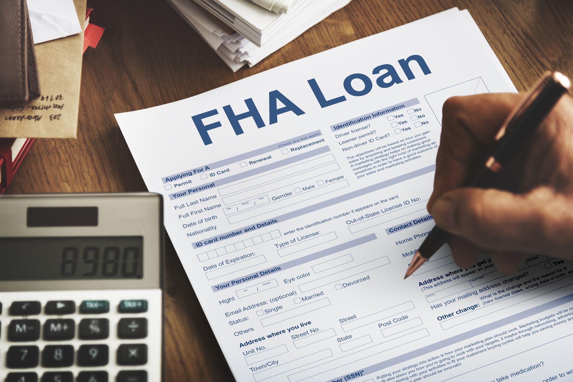 Person's hand filling out an FHA loan application form with a calculator on a wooden desk