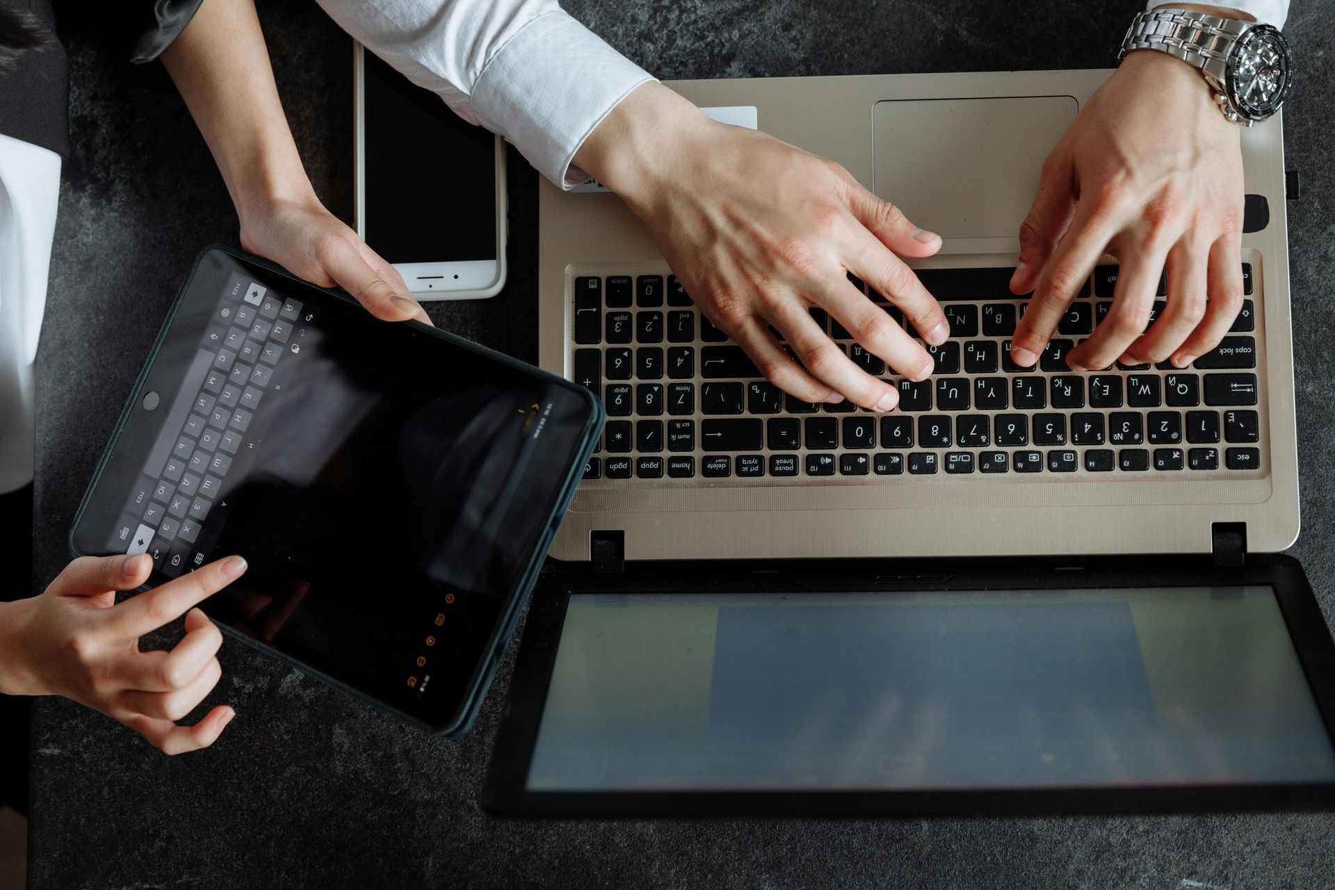 Two people work at a desk, one typing on a laptop and the other operating a tablet next to a smartphone. Two people work at a desk, one typing on a laptop and the other operating a tablet next to a smartphone.