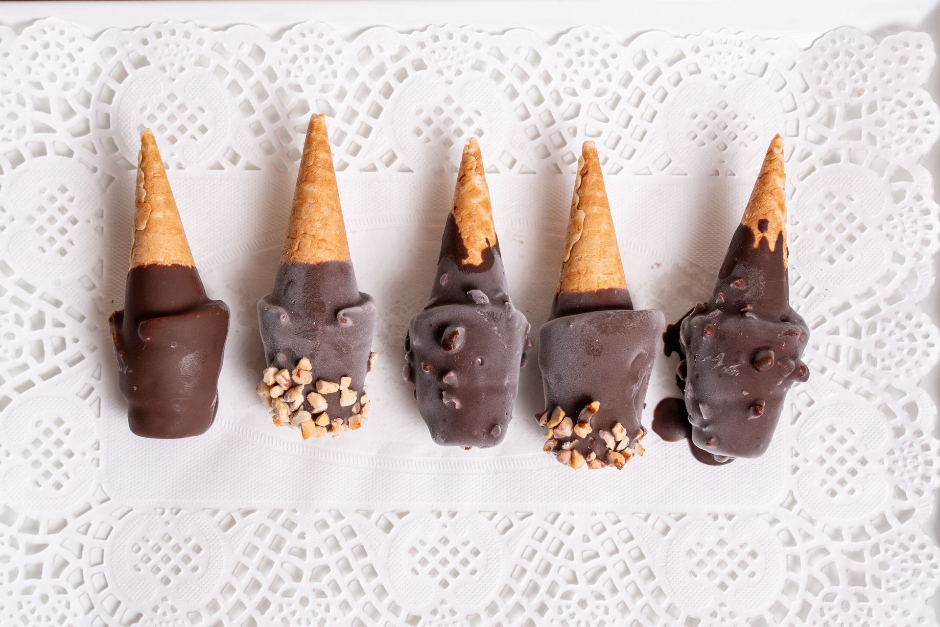 Cinque dolcetti a forma di cono ricoperti di cioccolato su uno sfondo di centrino di pizzo bianco.