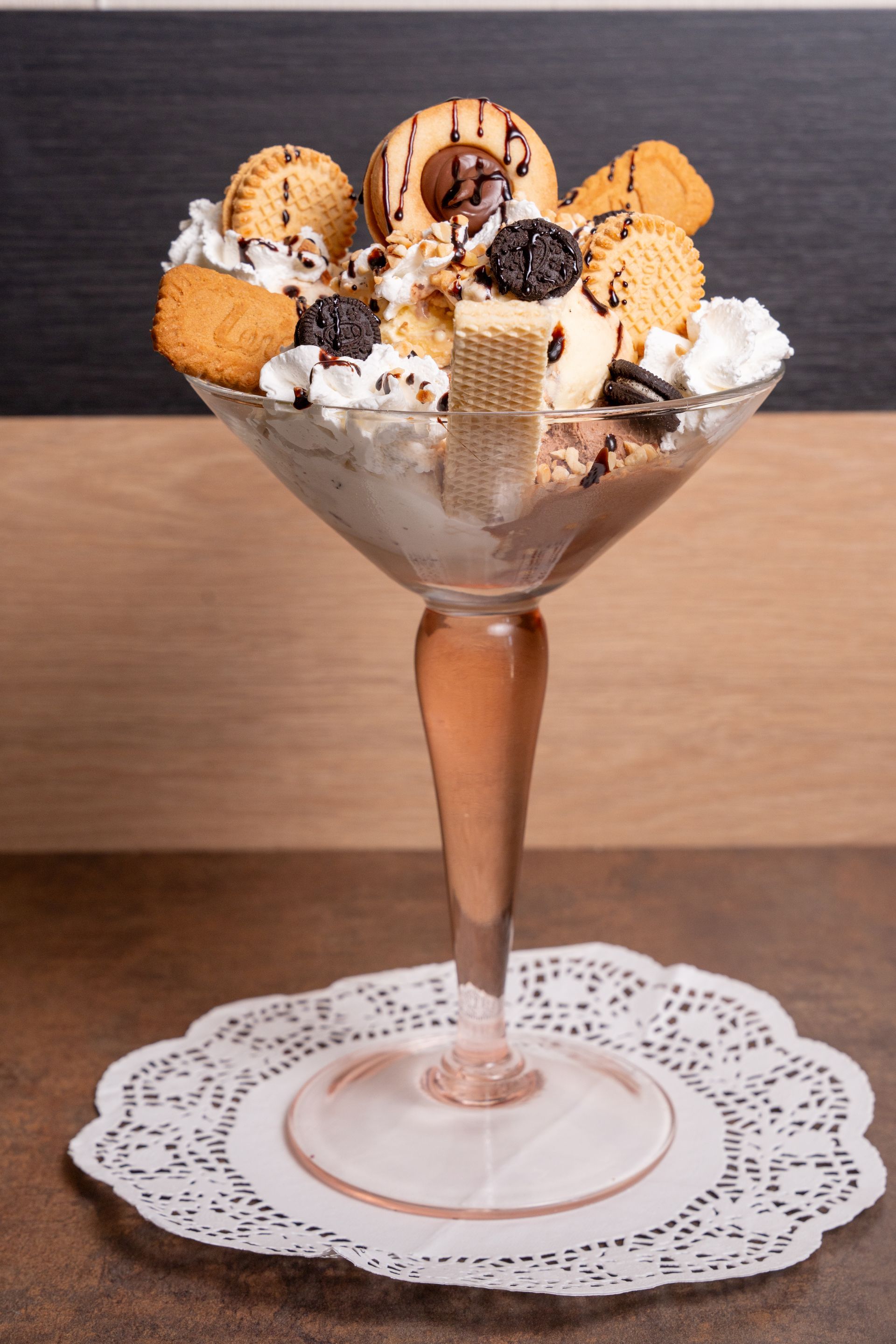 Coppa gelato in un bicchiere trasparente con stelo, guarnita con biscotti, cialde e panna montata su un centrino di pizzo.