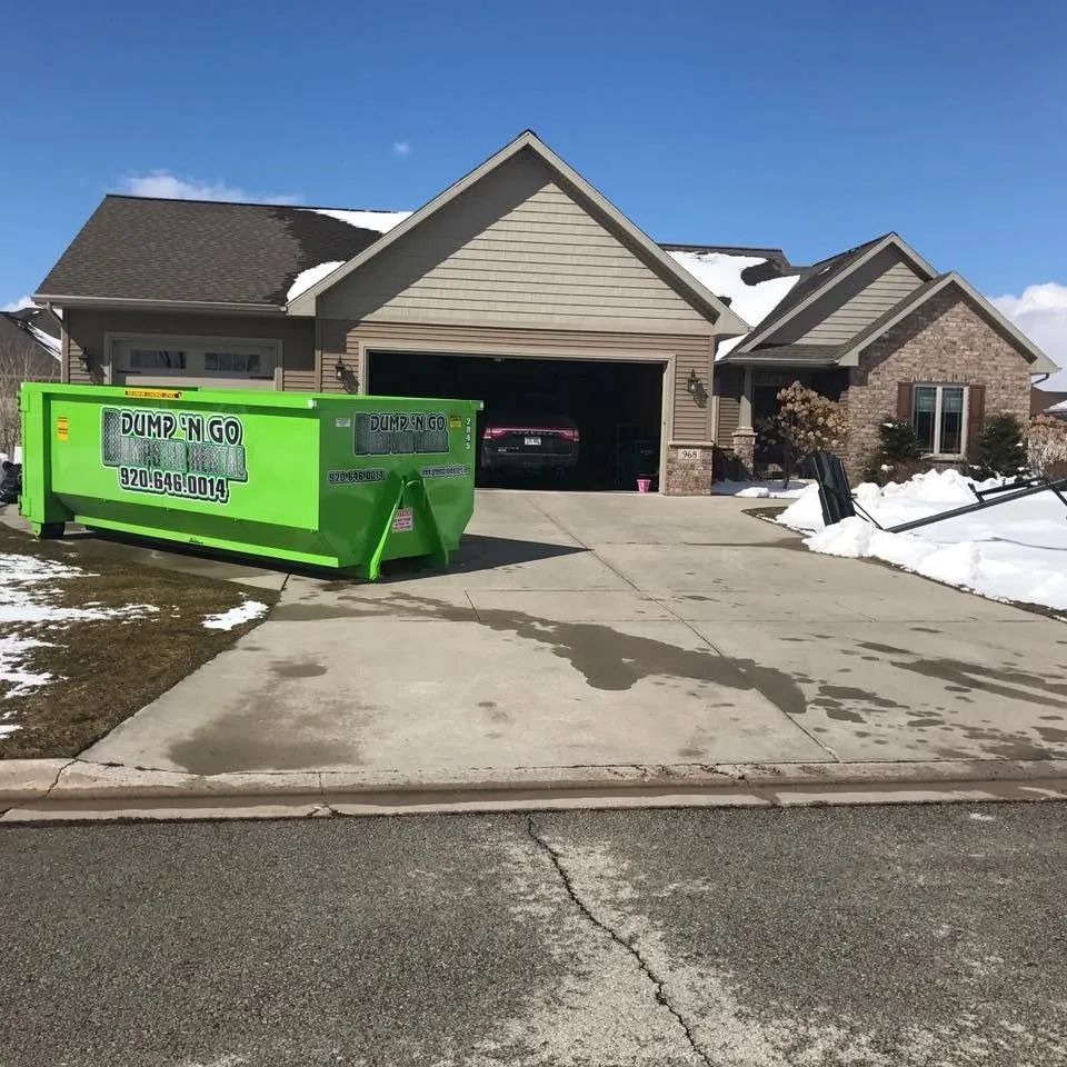 Bright green dumpster in a snowy suburban driveway in front of a house with an open garage