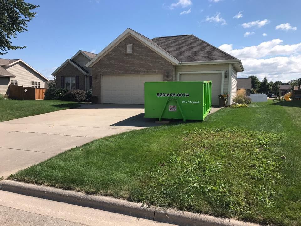 Green dumpster on a suburban driveway in front of a beige house on a sunny day