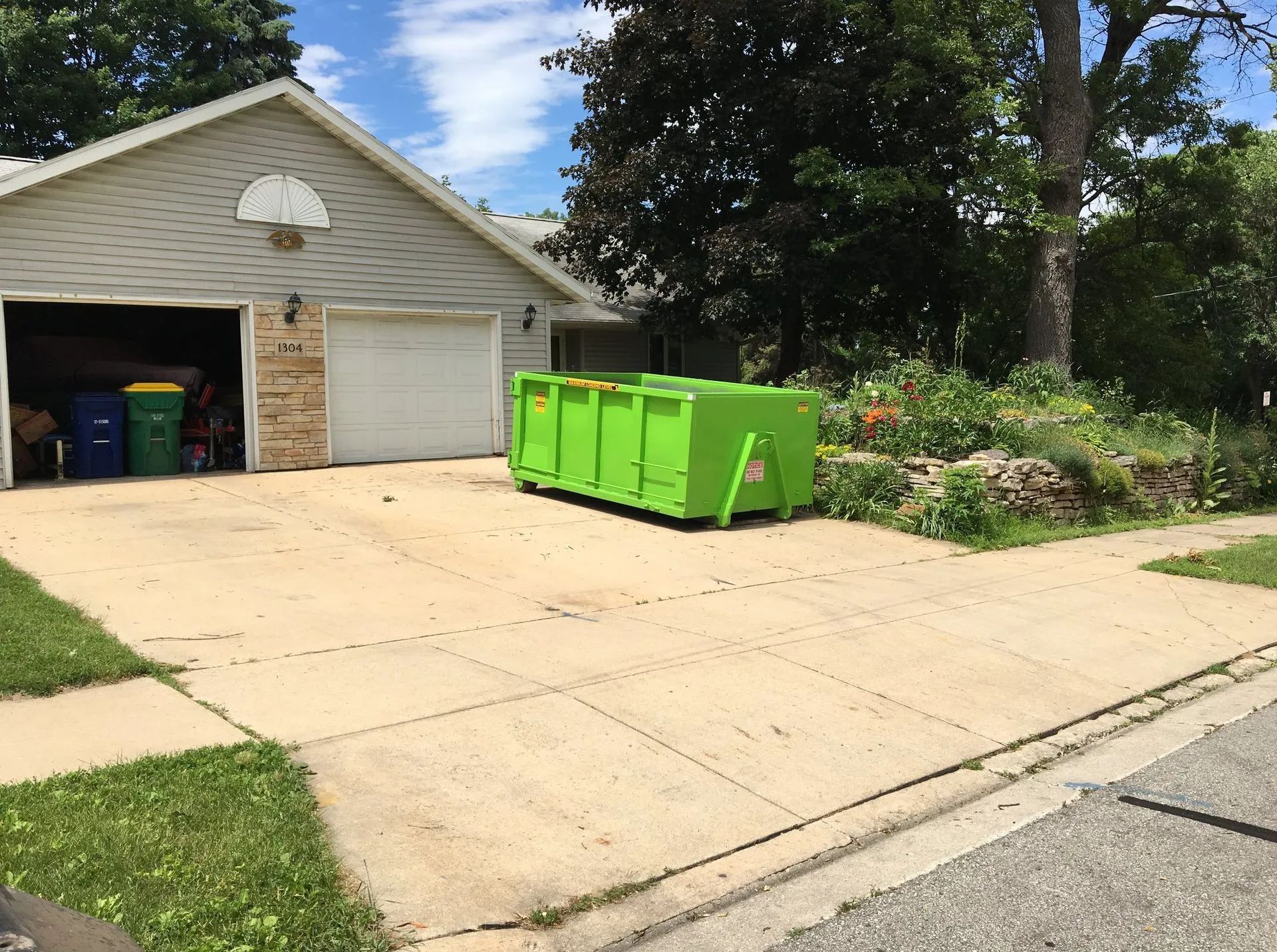 Green dumpster on a driveway in front of a garage, with a garden bed beside it.