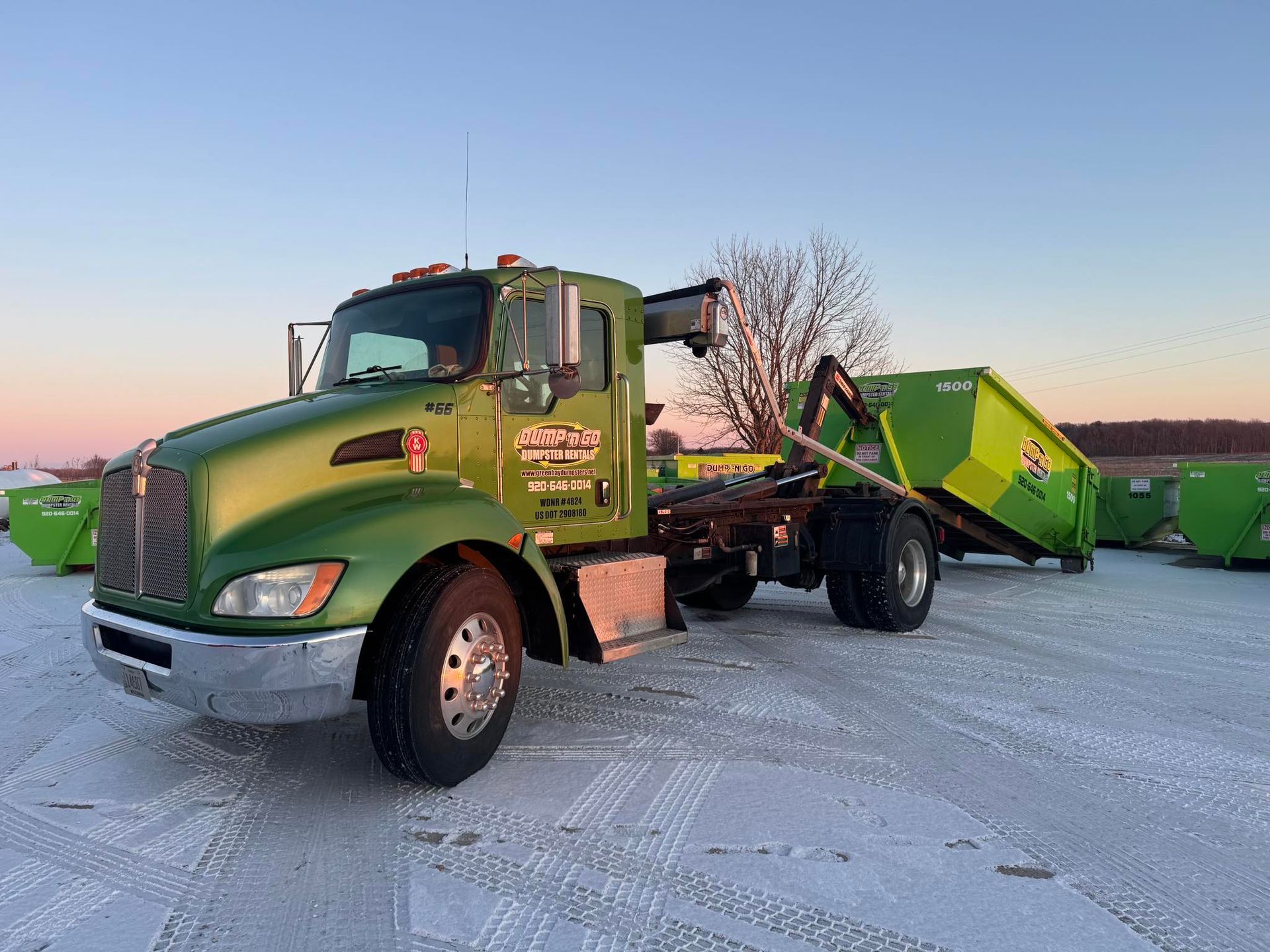 Green semi truck with a lime-green dumpster on a snowy lot at sunset
