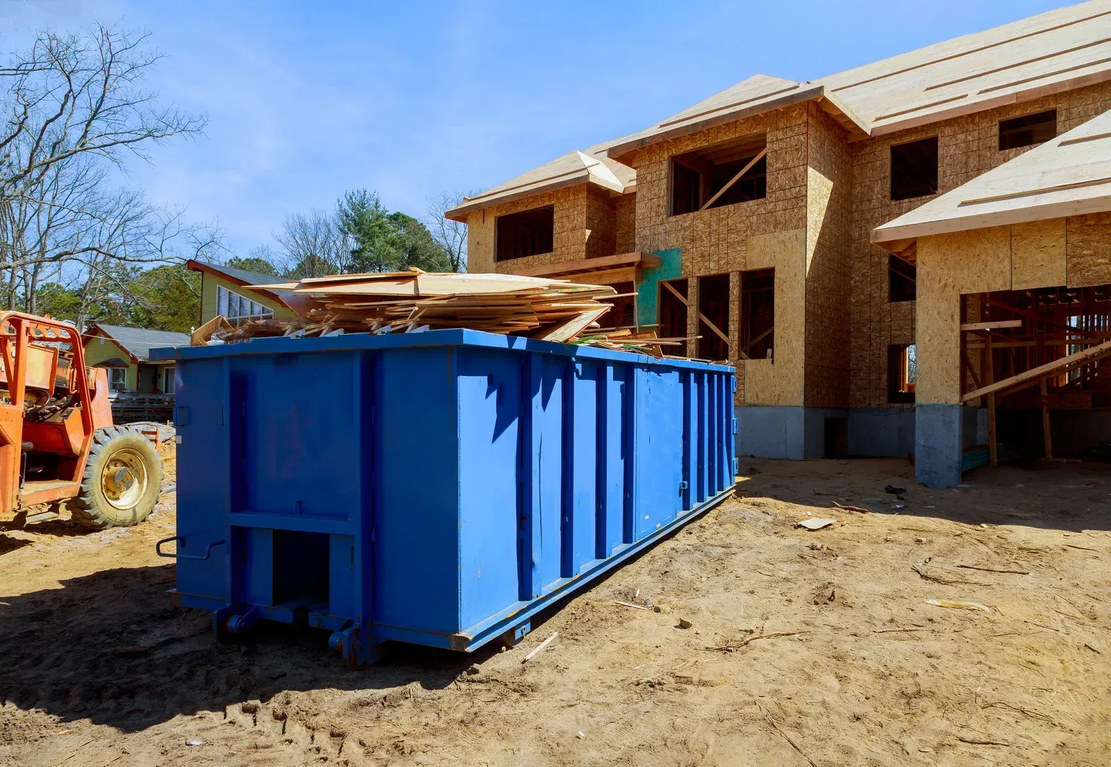 A blue dumpster sits in front of a new house under construction, with wooden boards piled on top of the container.