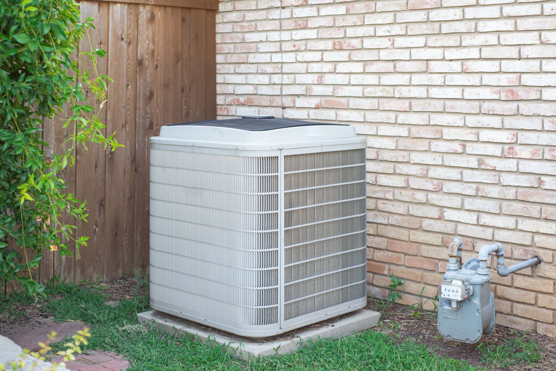Residential AC condenser at a brick wall surrounded by grass, a stone path, and utility piping Residential AC condenser at a brick wall surrounded by grass, a stone path, and utility piping