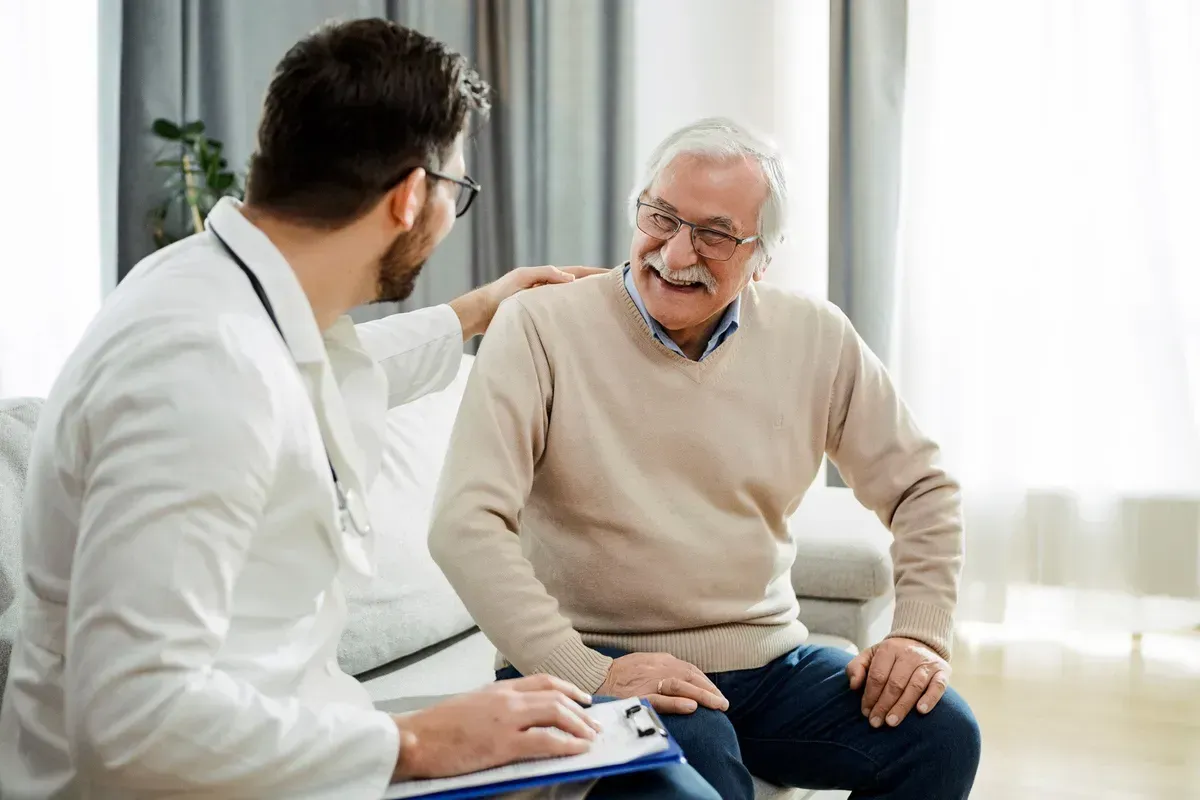 An elderly man is sitting on a couch talking to a doctor. An elderly man is sitting on a couch talking to a doctor.
