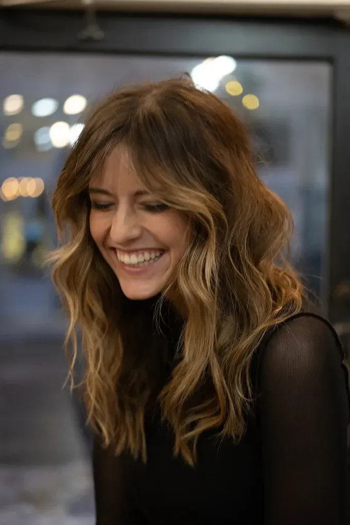 Woman with wavy brown and blonde hair smiles, wearing a black turtleneck.