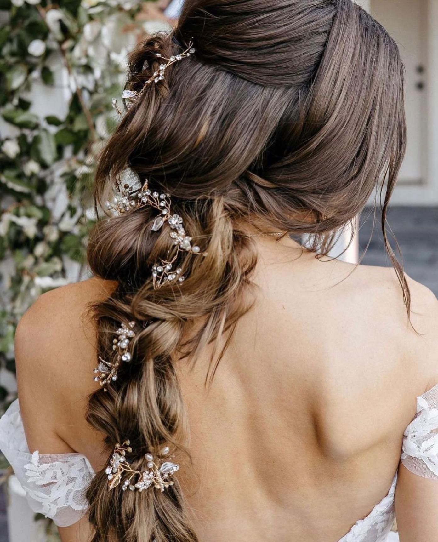 Woman with brown hair in a side braid hairstyle, embellished with jewels, wearing a white dress.