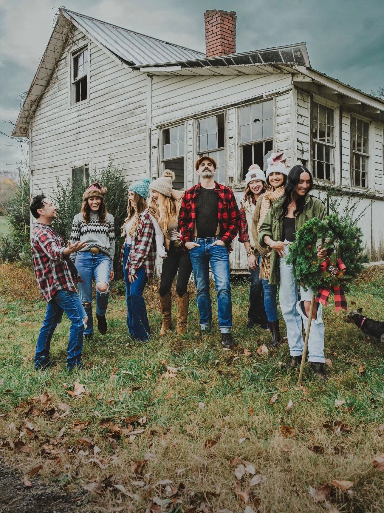 Group of people by a weathered house; some wear holiday hats, a person holds a wreath. Cloudy sky.