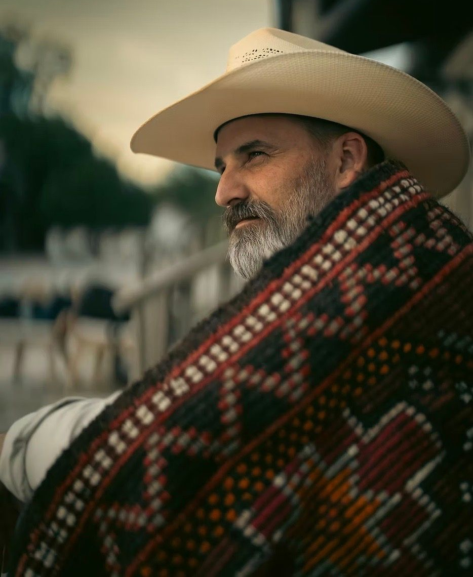Man in cowboy hat, wrapped in a patterned blanket, looking off to the side. Outdoor setting, muted colors.