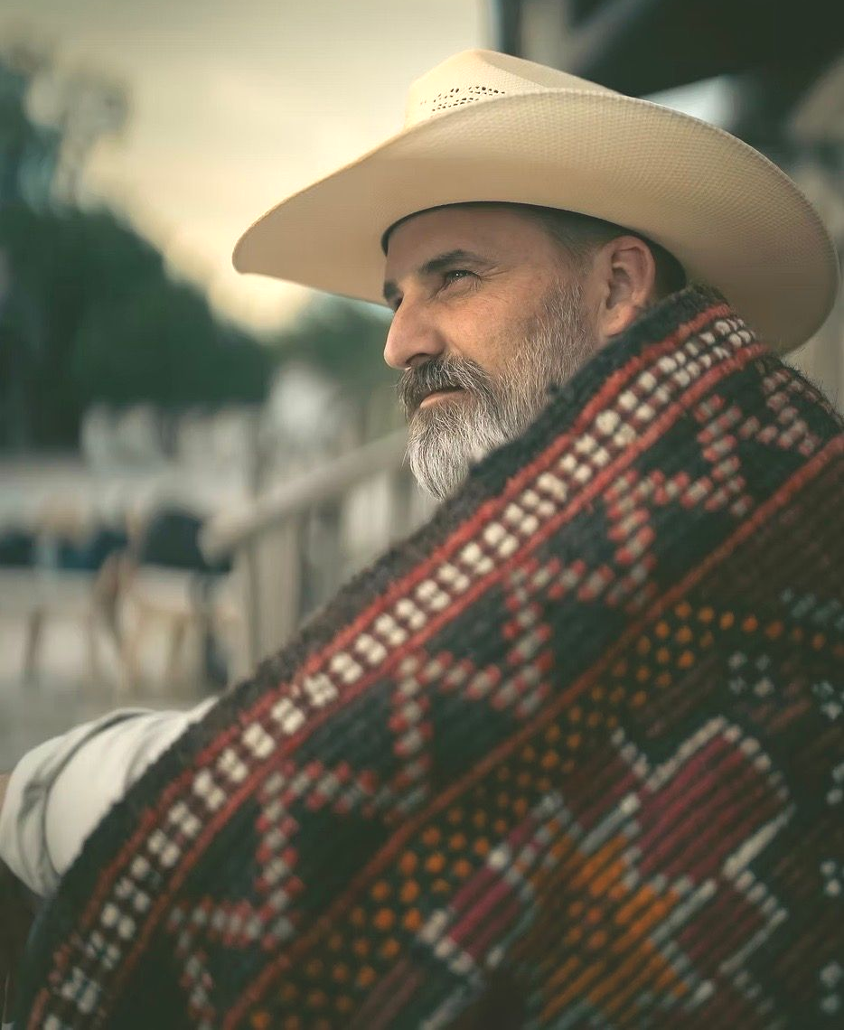 Man in cowboy hat, wrapped in a patterned blanket, looking off to the side. Outdoor setting, muted colors.