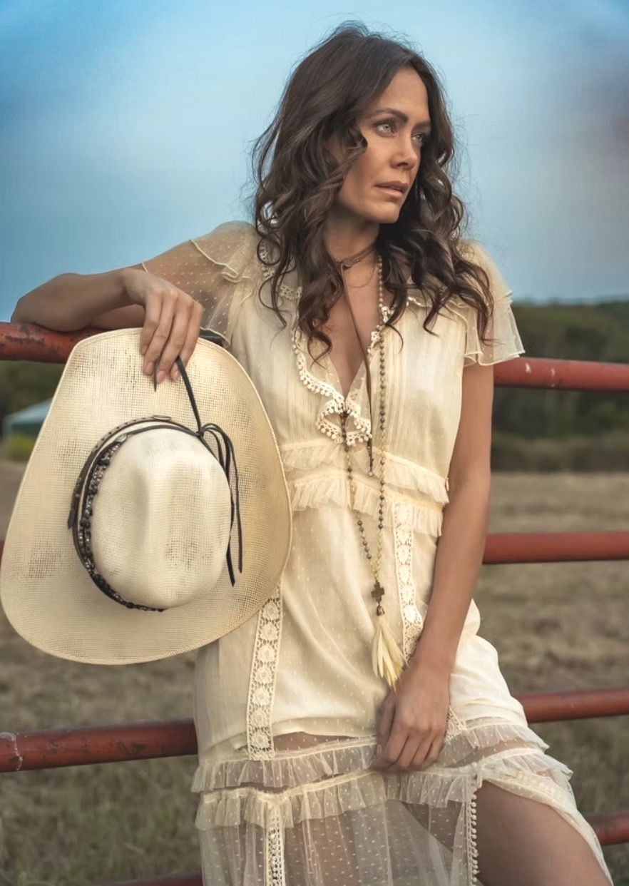 Woman in white dress leans on red fence, holding cowboy hat. Outdoors, thoughtful expression.