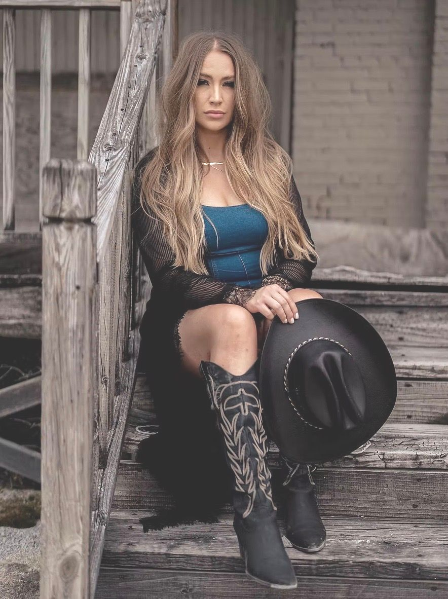 Woman in cowboy boots and hat sits on weathered steps, long blonde hair, blue top, black lace cover.