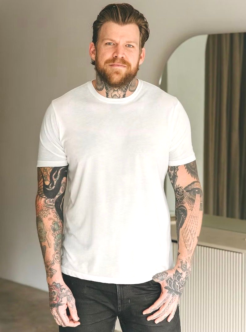 Man with tattoos wearing a white t-shirt and dark jeans, standing indoors with a neutral background.