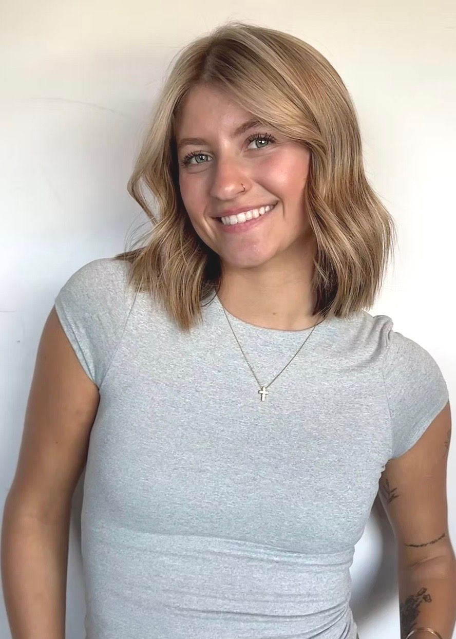 Smiling woman with shoulder-length blonde hair in a light gray T-shirt against a plain wall