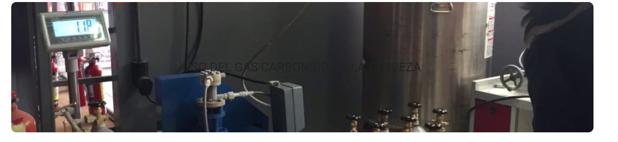 Equipo industrial con un monitor, maquinaria azul y grandes tanques de metal en un entorno de fábrica.