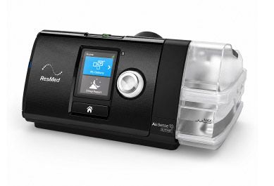 Máquina CPAP negra con pantalla pequeña y cámara humidificadora transparente a la derecha.