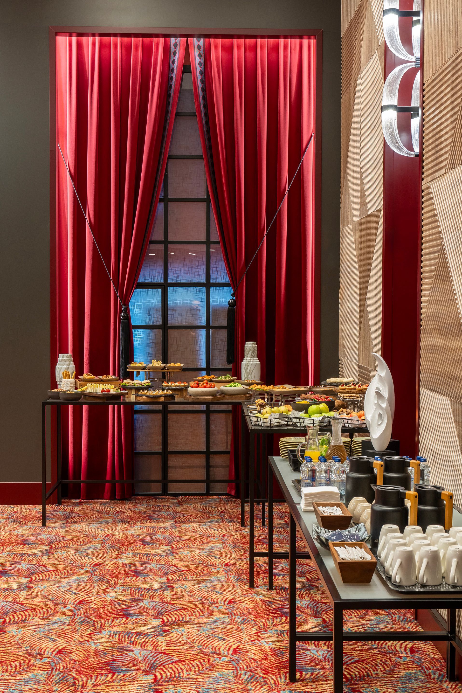 Elegant buffet setup with red curtains, window backdrop, and trays of food and drinks on black tables.