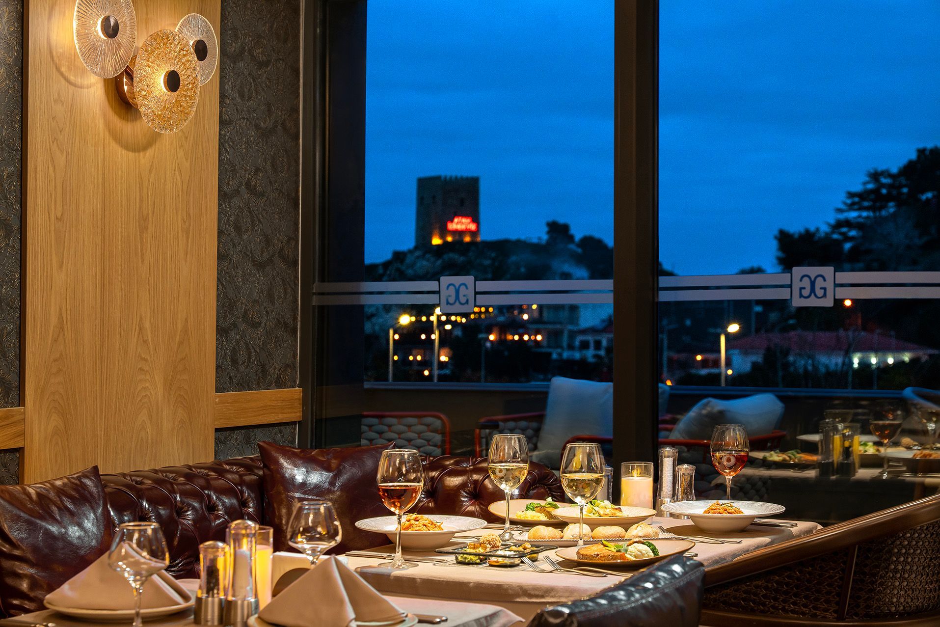 Elegant restaurant dining room with leather seating and candlelit table overlooking a blue cityscape at dusk