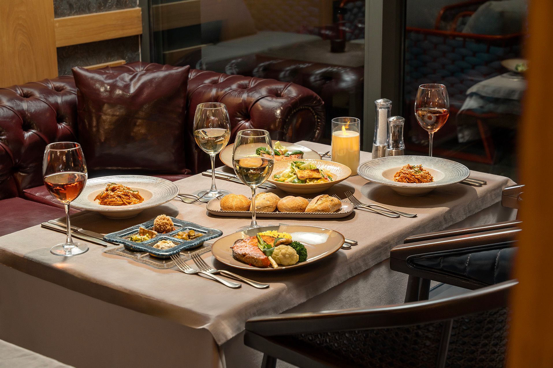 Restaurant table set with plated meals, glasses of wine, and a candle-lit leather booth by a window