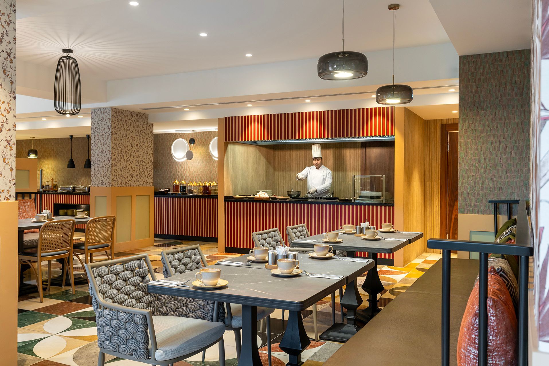 Hotel restaurant dining area with patterned chairs, set tables, and a chef at a buffet counter
