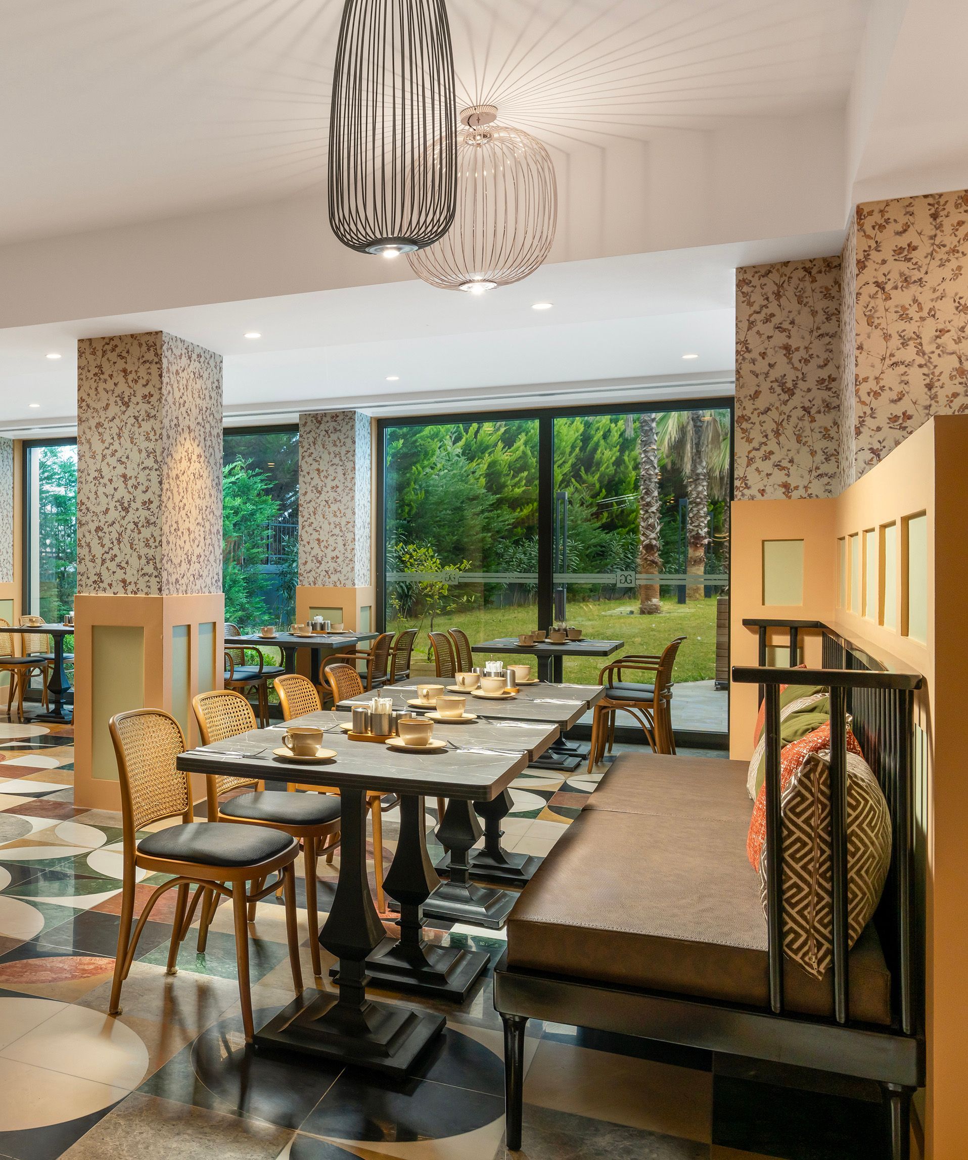 Bright dining room with wooden tables, patterned columns, and large windows overlooking a green garden